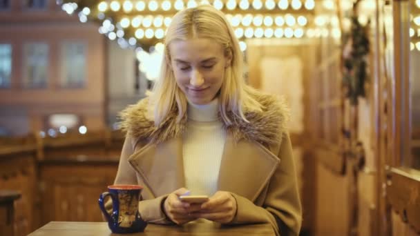 在圣诞市场上使用智能手机的妇女 女孩享受寒假季节 社交网络的概念 使用应用程序 背景上模糊的圣诞灯 — 图库视频影像