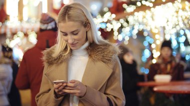 Noel pazarı üzerinde akıllı telefon kullanan kadın. Kız kış zevk tatil sezonu. İletişim, arka planda uygulama bulanık Noel ışıkları kullanarak sosyal ağ kavramı.