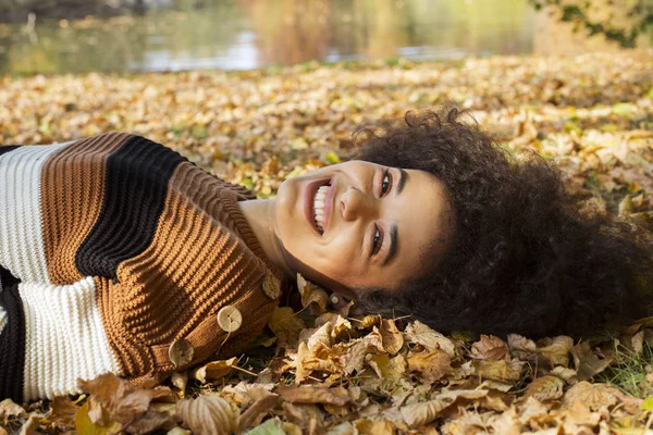 Güneşli parkta sonbahar yaprakları üzerinde uzanan genç Afro-Amerikan kadın..