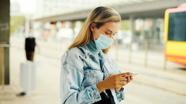 Maskeli genç sarışın bir kadın otobüs durağında bekliyor ve telefon kullanıyor..