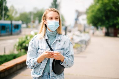 Antibakteriyel maske takan genç bir kadın şehirde telefon kullanıyor..