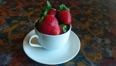 Renkli bir masa örtüsü üzerinde beyaz bir fincanın üzerinde çilekler.