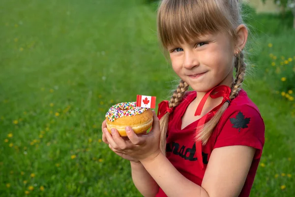「加拿大快乐日庆祝概念」。微笑的女孩拿着一个撒满了加拿大国旗的甜甜圈。年轻的加拿大小孩. 图库照片