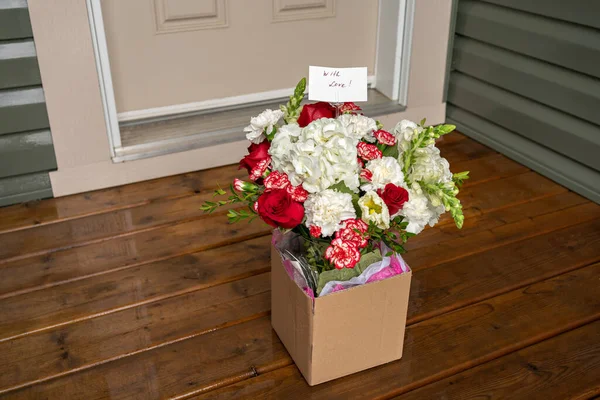 A bouquet of red white flowers in a carton box on a porch doorstep of a house. Surprise contactless delivery of flowers.