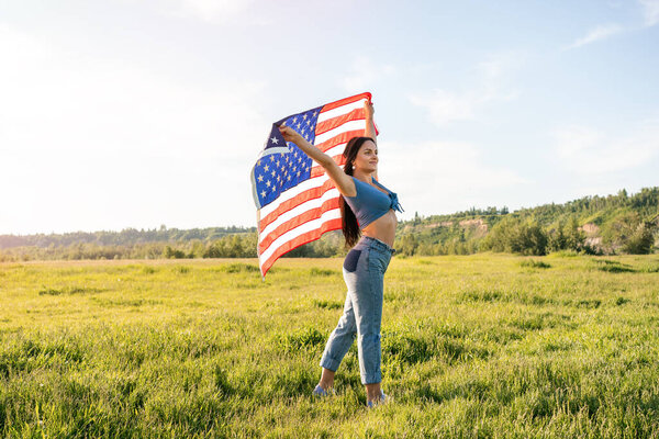 Woman wearing jeans and blue top holding an American flag above her head on the park field during sunset. Side view. 