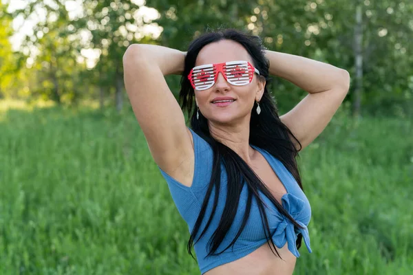 Happy Canada Day Celebration concept. Brunette woman with Canadian flag and themed sunglasses on nature background.