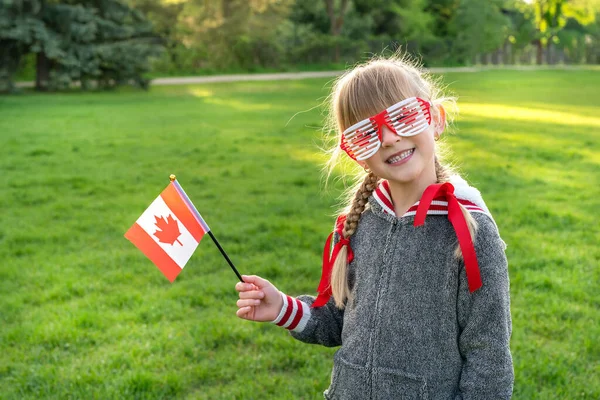 祝你佳节快乐！可爱的小女孩，拿着加拿大国旗，戴着主题太阳镜在公园里 . 免版税图库图片