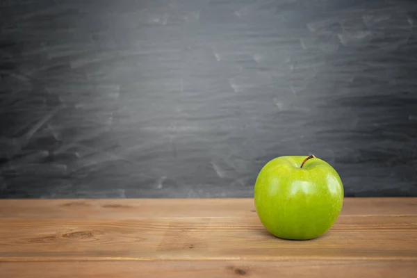 Green apple is on a wooden table and chalk board on the back with copy space as back to school or educational concept.