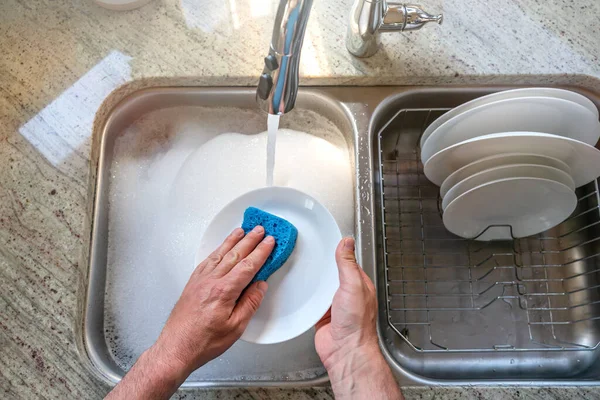 Ein Mann wäscht einen weißen Teller mit einem blauen Schaumschwamm in der Küche, von oben betrachtet. Geschirrspülen, Hausroutine, Tellerputzen. — Stockfoto