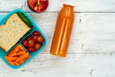 Okul yemeği - içinde sandviç, domates, havuç olan plastik kutu, beyaz ahşap arka planda portakal suyu olan bölümler..
