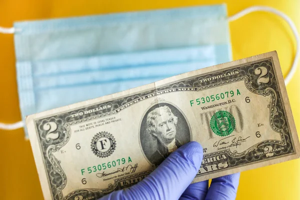 Lateks eldivendeki bir el, tıbbi bir maskenin arka planında iki dolar tutar. Yeni Coronavirüs salgınının küresel ekonomi ve mali kriz üzerindeki küresel etkisi.