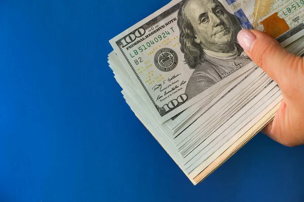 A wad of hundred-dollar bills held in his hand against a bright blue background. The concept of cash, give money on credit, pay in cash