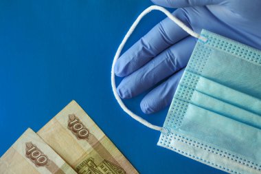 Eldivenli eller tıbbi maskelerin arka planında 100 ruble tutuyor. Salgın sırasında kişisel koruyucu ekipman maliyetinin azaltılması ve azaltılması.