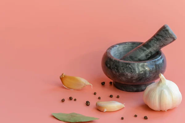 A marble mortar and pestle next to an artistically scattered whole black pepper, a Bay leaf and a garlic bulb on a pink background. Spices