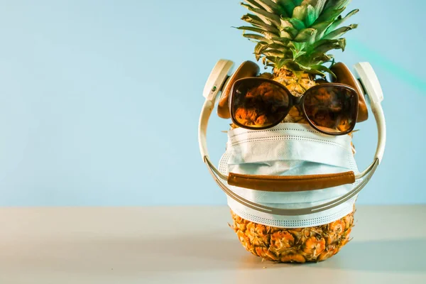 Güneş gözlüğü, kulaklık ve tıbbi maske takan yaratıcı olgun ananas. Salgın sırasında yaz tatili kavramı. Boş alan