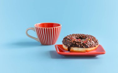 Taze, çikolata kaplı bir donut dikdörtgen kırmızı bir tabakta mavi arka planda kırmızı çizgileri olan beyaz bir porselen kupanın yanında oturur. Tatlı yemek..