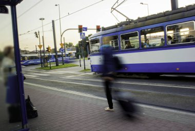 Krakow şehrinde yoğun bir hayat manzarası ve tramvaydan önce evrak çantasıyla yürüyen bir adamın uzun pozlu görüntüsü.