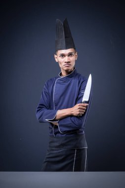 Mavi üniformalı, siyah önlüklü, koyu gri arka planda siyah şapkalı ve bıçaklı Asyalı bir yönetici şefin portresi.