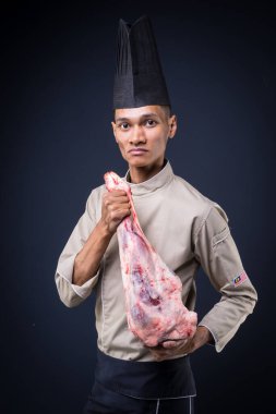 Asyalı bir yönetici şefin portresi açık kahverengi üniformalı, siyah önlüklü ve gri arka planda siyah şapkalı kuzu budu etli..