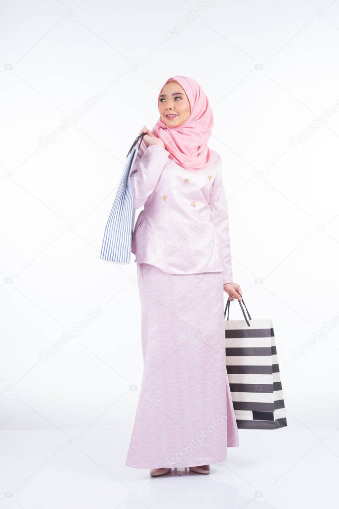 A beautiful Muslim female model in a Asian traditional dress carrying shopping bags isolated on white background. Idul fitri festive preparation shopping concept. Full length portrait.