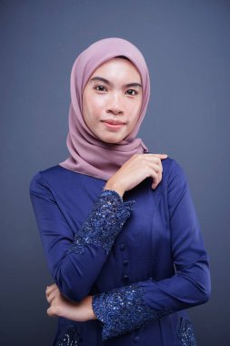Koyu mavi, tesettürlü bir modern kebaya giyen güzel bir Müslüman kadın model. Gri arka planda izole edilmiş Asyalı Müslüman geleneksel bir elbise. Eidul Fitri moda ve yaşam tarzı portresi konsepti.