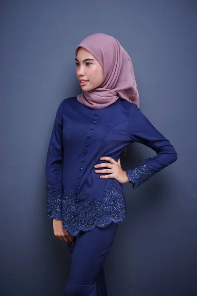 A beautiful Muslim female model wearing a dark blue modern kebaya with hijab, an Asian Muslim traditional dress isolated on grey background. Eidul fitri fashion and lifestyle portrait concept.