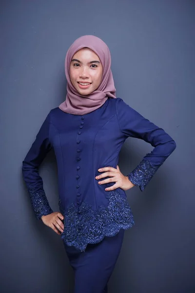 A beautiful Muslim female model wearing a dark blue modern kebaya with hijab, an Asian Muslim traditional dress isolated on grey background. Eidul fitri fashion and lifestyle portrait concept.