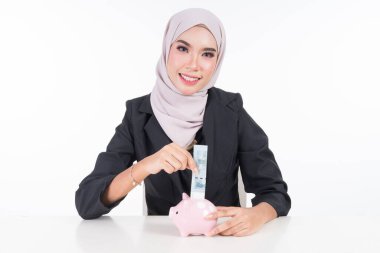 Finans ve muhasebe tasarruf kavramı. İş dünyasından Müslüman bir kadın beyaz arka planda izole bir şekilde kumbaraya bir banknot yerleştiriyor..