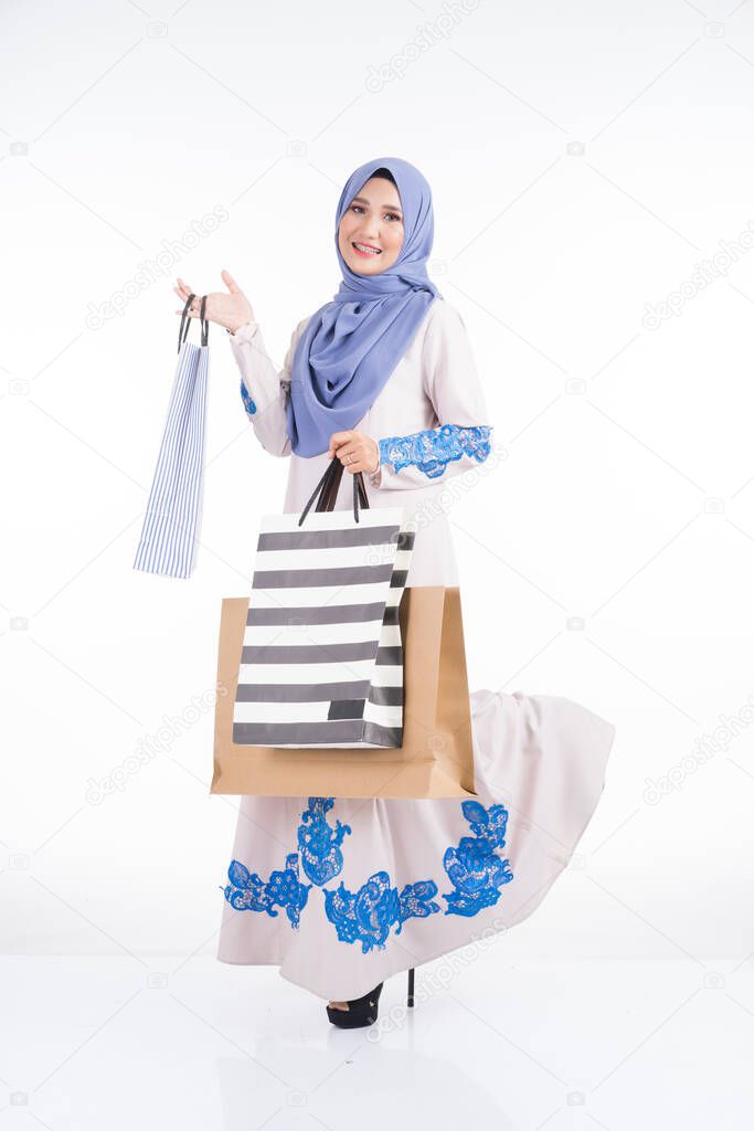A beautiful Muslim female model in a Asian traditional jubah dress carrying shopping bags isolated on white background. Eidul fitri festive shopping concept. Full length portrait.