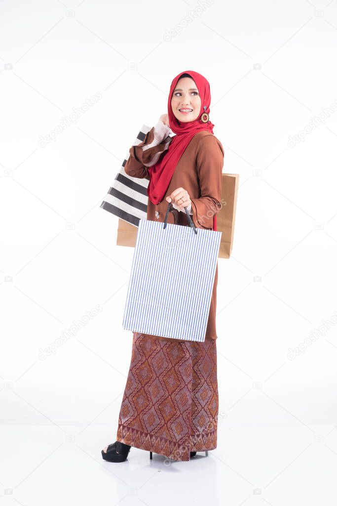 A beautiful Muslim female model in a Asian traditional dress modern kebaya carrying shopping bags isolated on white background. Eidul fitri festive preparation shopping concept. Full length portrait.