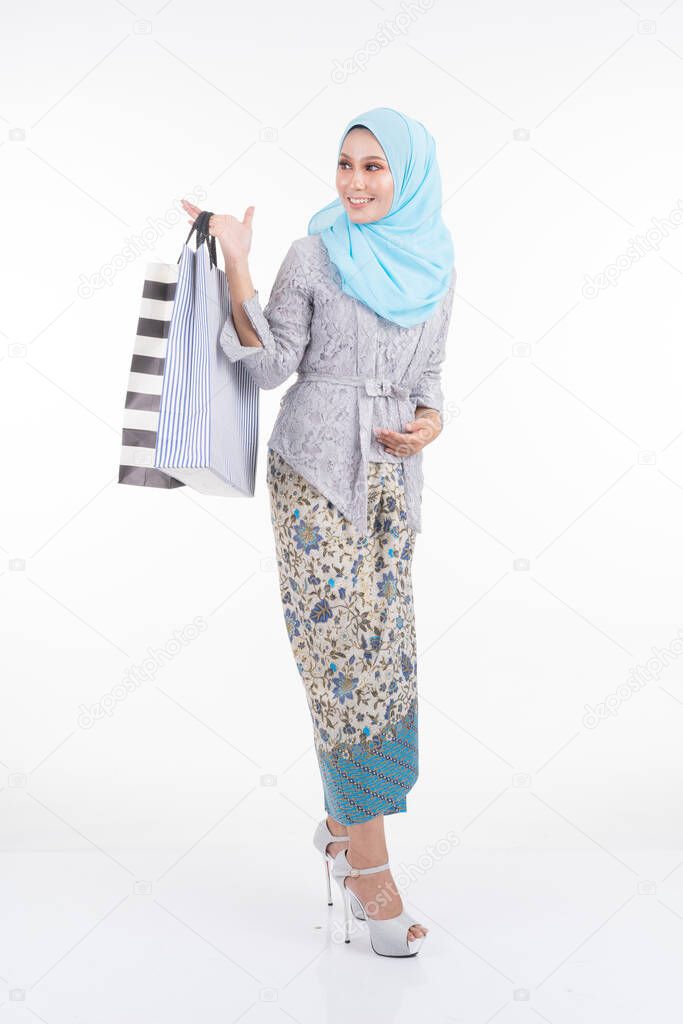 A beautiful Muslim female model in a Asian traditional dress modern kebaya and hijab carrying shopping bags isolated on white background. Eidul fitri fashion and festive shopping concept