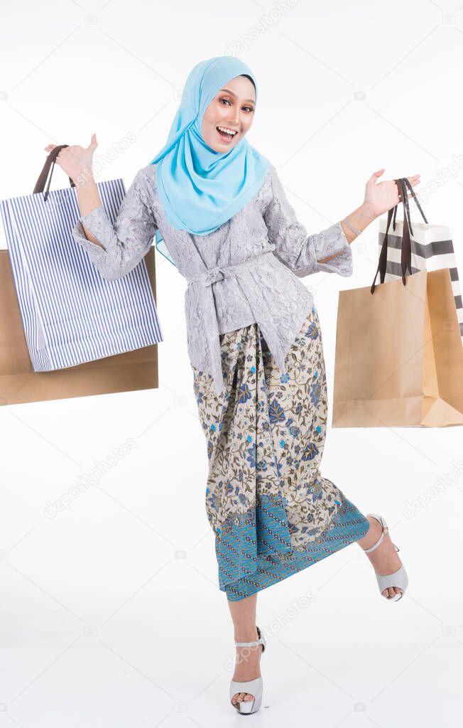 A beautiful Muslim female model in a Asian traditional dress modern kebaya and hijab carrying shopping bags isolated on white background. Eidul fitri fashion and festive shopping concept