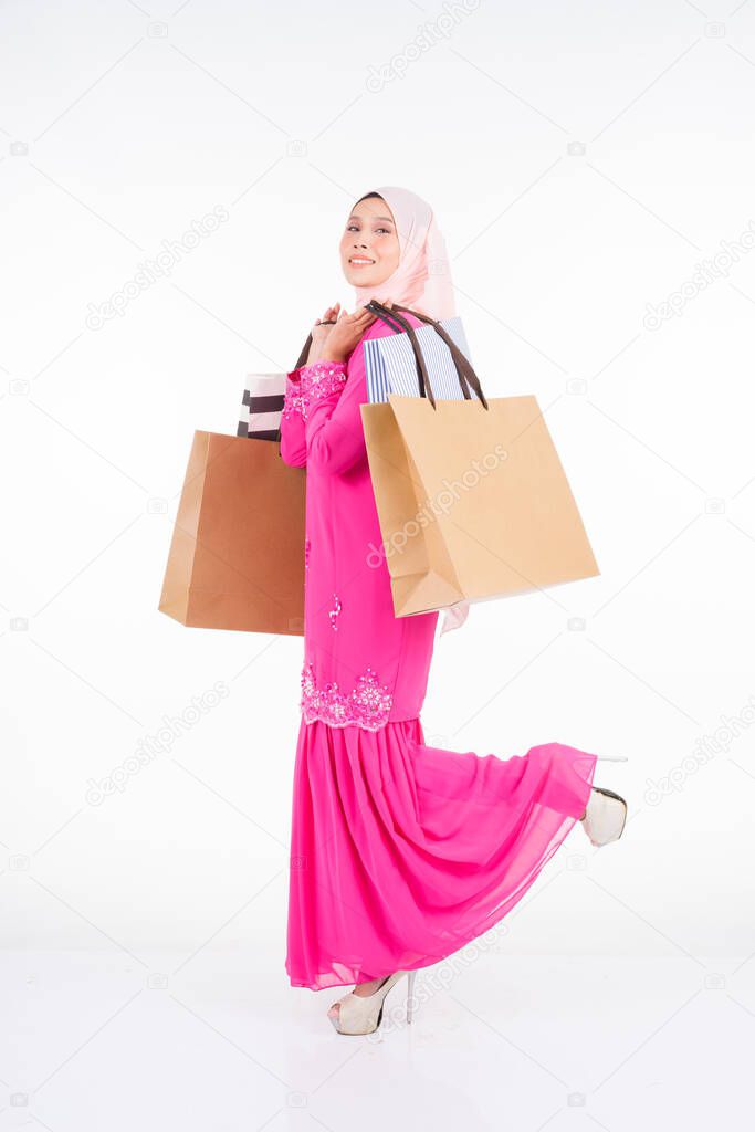 A beautiful and excited Muslim female model in a traditional pink modern kurung carrying shopping bags isolated on white background. Eidul fitri fashion and festive shopping concept