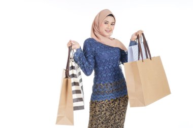 Geleneksel Asya elbisesi içinde güzel ve heyecanlı bir kadın model beyaz arka planda izole edilmiş alışveriş torbaları taşıyan modern bir kurung. Eidul Fitri moda ve festival hazırlık alışveriş konsepti.