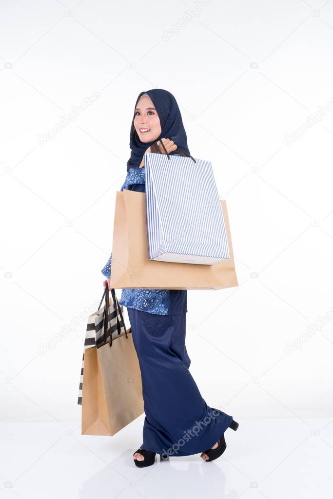 A beautiful and excited Muslim female model in a Asian traditional dress modern kurung carrying shopping bags isolated on white background. Eidul fitri fashion and festive preparation shopping concept