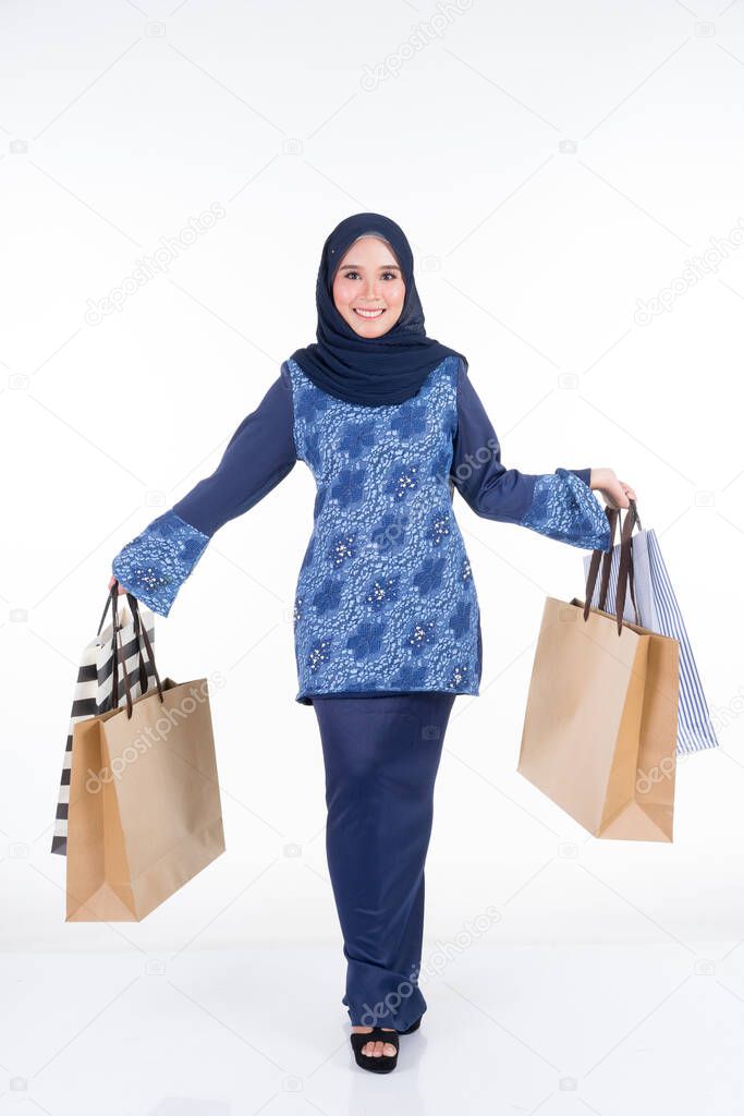 A beautiful and excited Muslim female model in a Asian traditional dress modern kurung carrying shopping bags isolated on white background. Eidul fitri fashion and festive preparation shopping concept