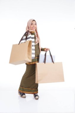 Geleneksel Asya elbisesi içinde güzel bir Müslüman bayan üniversite öğrencisi beyaz arka planda izole edilmiş alışveriş torbaları taşıyan modern kurung. Eidul Fitri moda ve festival alışveriş konsepti.