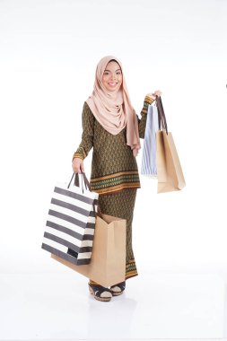 Geleneksel Asya elbisesi içinde güzel bir Müslüman bayan üniversite öğrencisi beyaz arka planda izole edilmiş alışveriş torbaları taşıyan modern kurung. Eidul Fitri moda ve festival alışveriş konsepti.