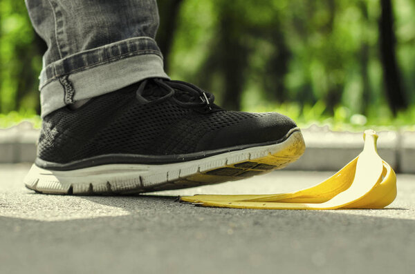 Men's foot in saturated black trainers slides on a yellow peel from banana on the background of summer park.