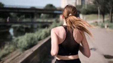 Şehirde nehir kenarında koşan genç bir kadın. Arkadan koşan bir kadının görüntüsü. Yolda koşan enerjik bir kadın