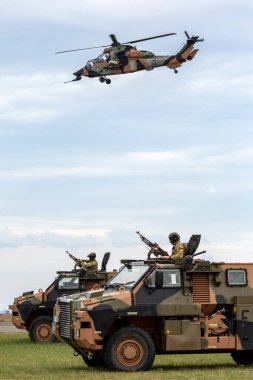 Avalon, Australia - February 27, 2015: Two Australian Army Bushmaster armoured Personnel carriers (APC) with an Army Eurocopter Tiger helicopter providing air cover.  clipart