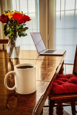 A coffee cup, a computer, floweres, all sitting on a table. clipart