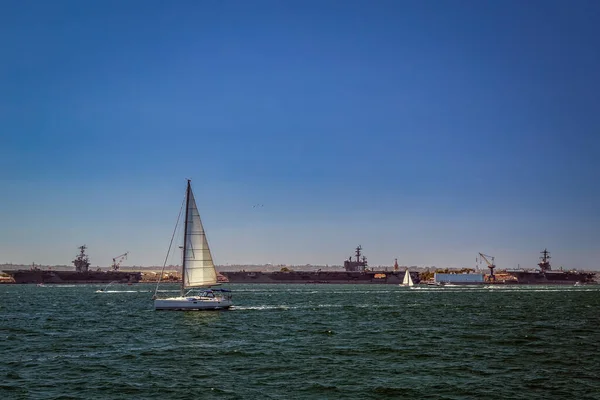A sailboat passing by three US Navy aircraft carriers dock at Coronado in San Diego Bay, California.