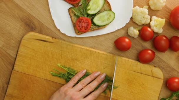 トップビュー女性の手は まな板にナイフでディルをカット キュウリとサンドイッチにそれを置く トマト アルグラと全小麦ライ麦パン ビーガンランチを作る女性 木のテーブルの上の新鮮な農場の野菜 — ストック動画