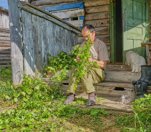 Senior Asian man ties birch brooms for a bath sitting on a wooden porch of an old country house in a Siberian village, Russia