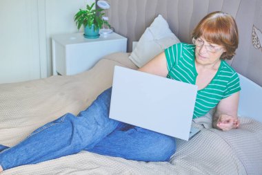 Üst düzey bir kadın evdeki yatak odasında dizüstü bilgisayar kullanıyor. Mutlu emekli insan yatakta online alışveriş yapıyor. Yaşam tarzı. Boşluğu kopyala