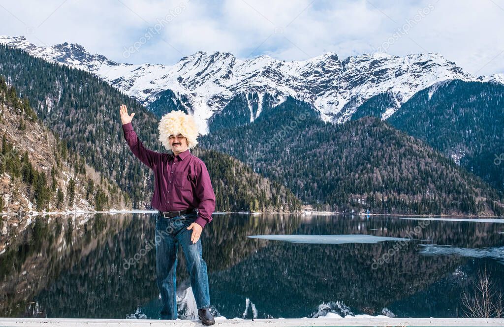 An Asian man in a traditional Caucasian white sheepskin hat, in a red shirt and jeans raised his hand in greeting as a backdrop of a beautiful mountain landscape and Lake Ritsa, Abkhazia.