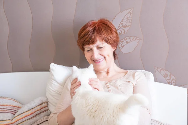 一位穿着家居服 红头发的快乐的老妇人坐在床上爱抚一只可爱的白猫 慢条斯理复制空间 — 图库照片