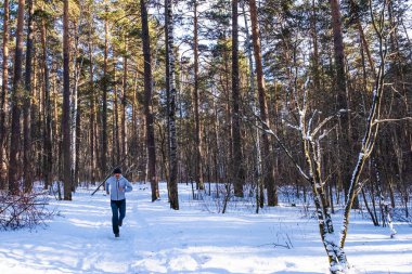 Açık renk bir kazak, kot pantolon ve örgü şapkalı bir adam güneşli bir günde, karlı bir ormanda karlı bir yolda koşar..