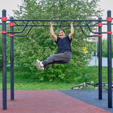 Mutlu kıdemli Asyalı adam parktaki çocuk parkında çiçek açan bir bitkinin arka planında antrenman yapıyor. Çapraz direkte asılı duruyor. Yaşam tarzı, sağlıklı alışkanlıklar. Hareket bulanıklığı